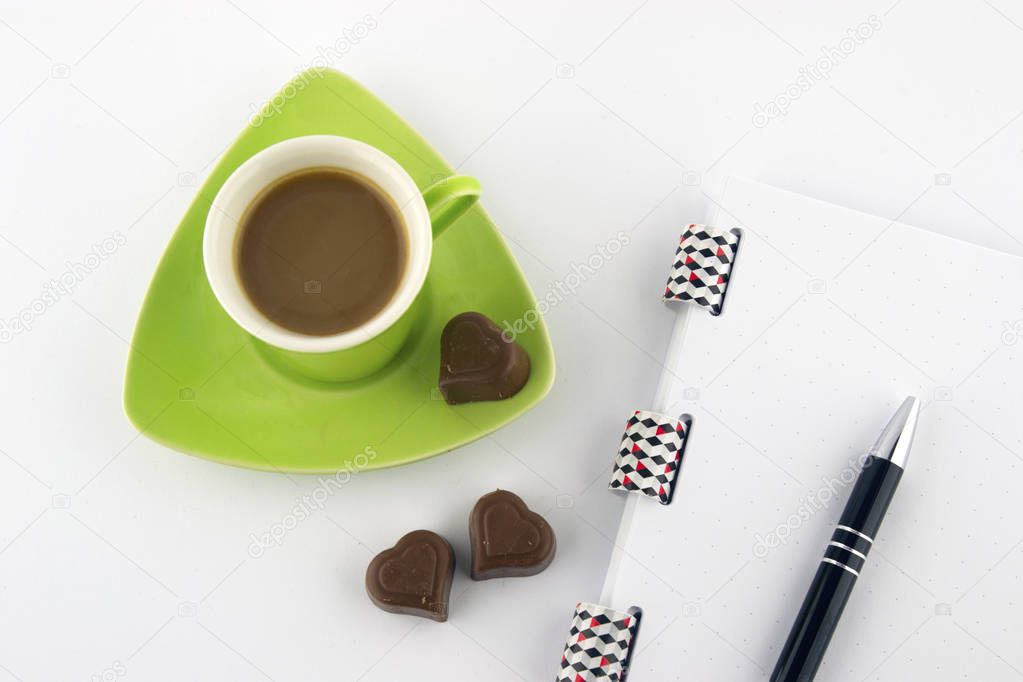 Coffee in a green cup with heart-shaped chocolate, notes and pencil on a white background. Top view.