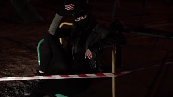 Девушка в черной маске вируса сидит за лентой забора смотрит в камеру — стоковое видео