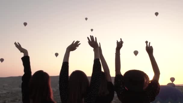 Cuatro niñas de pie frente a la puesta del sol y tirar de sus brazos hacia arriba en medio de un hermoso paisaje y globos — Vídeo de stock