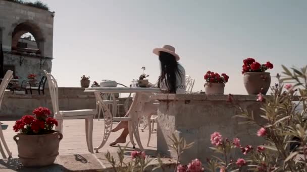 Jolie jeune brune à l'aspect européen dans une belle robe blanche aux lunettes élégantes noires et un chapeau assis à une table avec un carnet ouvert et profite du temps chaud — Video