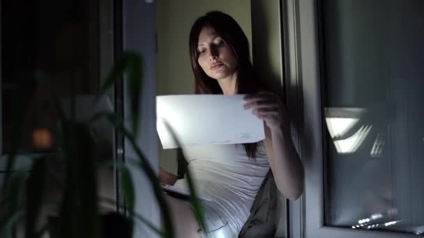Девушка в белой футболке и джинсовых шортах работает на ноутбуке поздно ночью на подоконнике — стоковое видео