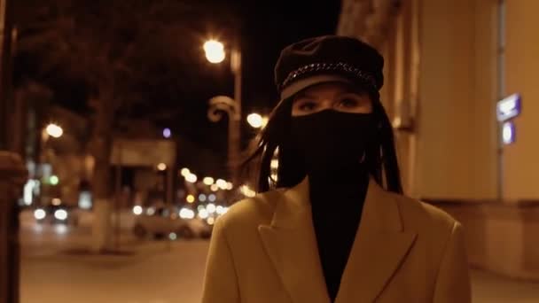 Dziewczyna w czarnym wirusie maska w beżowym płaszczu przechodzi przez ulicę w nocnym mieście — Wideo stockowe