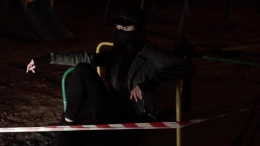 Siyah virüs maskeli kız çitin arkasında oturuyor ve kameraya bakıyor.