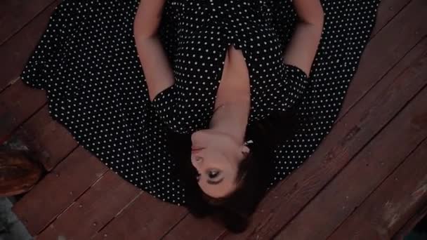 A beautiful girl of European appearance with black hair in a black dress with white polka dots lies on the wooden floor and pulls her hands to the camera — Stock Video