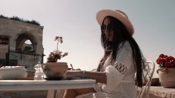 Hübsche junge Brünette mit europäischem Aussehen in einem schönen weißen Kleid mit schwarzer stylischer Brille und Hut sitzt an einem Tisch und schreibt, macht Notizen — Stockvideo
