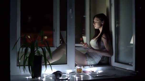 Chica en una camiseta blanca y pantalones cortos de mezclilla trabaja en un ordenador portátil tarde en la noche en el alféizar de la ventana — Vídeos de Stock