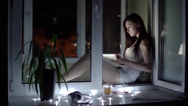 Chica en una camiseta blanca y pantalones cortos de mezclilla trabaja en un ordenador portátil tarde en la noche en el alféizar de la ventana — Vídeos de Stock