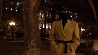 Siyah virüs maskeli, bej ceketli bir kız, gece parkında bir ağaçta duruyor.