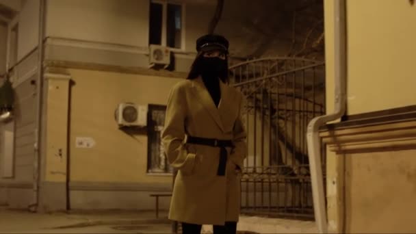 Ragazza in maschera virus nero in un cappotto beige passeggiate lungo edificio nella città di notte — Video Stock