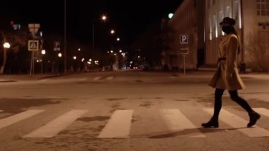 Siyah virüs maskeli, bej ceketli bir kız gece vakti karşıdan karşıya geçiyor.