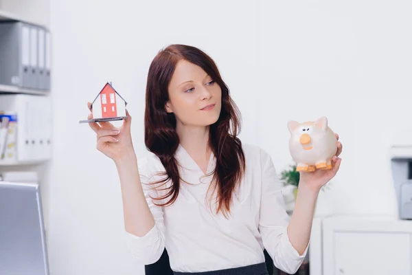 attractive woman holding little house model and piggybank in office