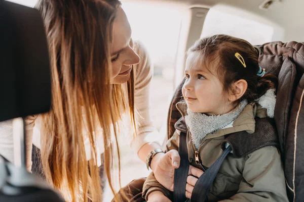 Mother securing her toddler daughter in the coat buckled into her baby car seat