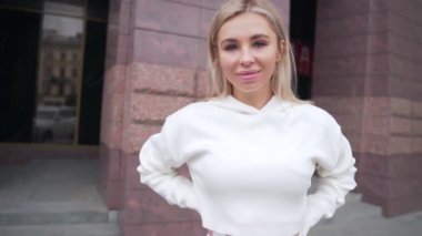 Beyaz eşofman giymiş, saçlarına dokunup düzenleyen çekici sarışın bir kız..