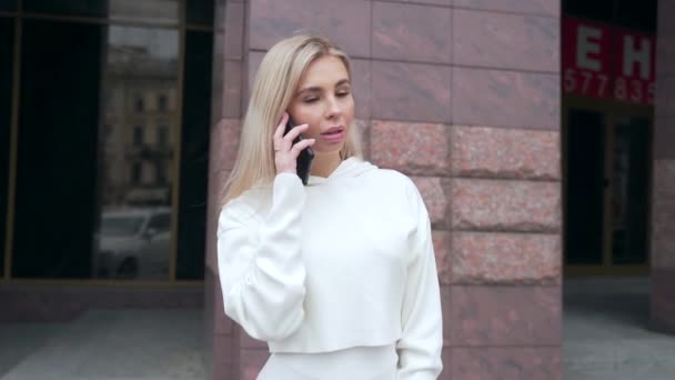 一个穿着白色西服的年轻漂亮的金发女人站在电话里笑着的画像 是在谈话 希斯特的女学生恋爱了城市街道背景 — 图库视频影像