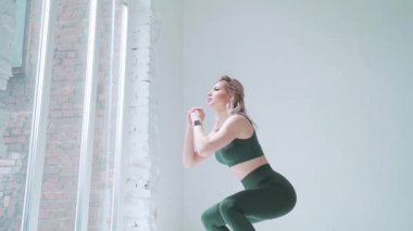 Seksi sarışın atletik vücut şekli. Spor hocası nefesiyle çömeliyor. Yeşil spor giyim taytları. Sağlam kalçalar. Pencereli beyaz bir arka plan duvarında. Stüdyo ya da salon. yavaş çekim