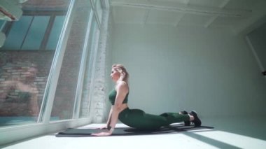 Geniş pencereli beyaz bir odada yoga yapan genç ve güzel bir kadın. Sağlık konsepti. Huzur ve huzur, kadın mutluluğu. Bir kişi. Seksi moda sarışın fitness eğitmeni