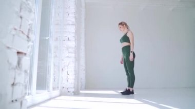 Atletik kadın beyaz duvara yaklaşıyor ve amuda kalkıyor. Denge ve spor, fitness yaşam tarzı. Beyaz arka plan stüdyosunda ya da geniş pencereli salonda yoga yapan genç bir kız.