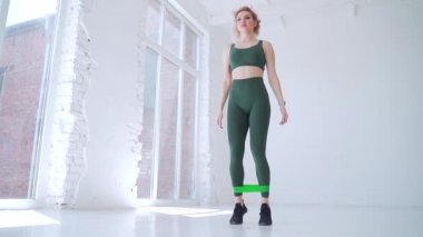 Genç bayan sporcu direniş bandıyla egzersiz yapıyor. Seksi sarışın fitness antrenörü lastik bantlarla çömelme hareketleri yapıyor. Pencereli beyaz bir arka plan duvarında. Stüdyo ya da salon. yavaş çekim