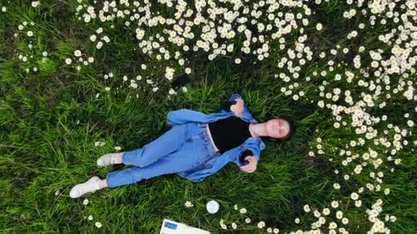 花の間の芝生の上に寝そべっている若いヒップスターの女の子はヘッドフォンリラックスで音楽を聴く 女子学生生活のリラックスと穏やかな 公園の芝生の上の静かな場所 空中からの眺め — ストック動画