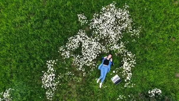 花の間の芝生の上に寝そべっている若いヒップスターの女の子はヘッドフォンリラックスで音楽を聴く 女子学生生活のリラックスと穏やかな 公園の芝生の上の静かな場所 空中からの眺め — ストック動画