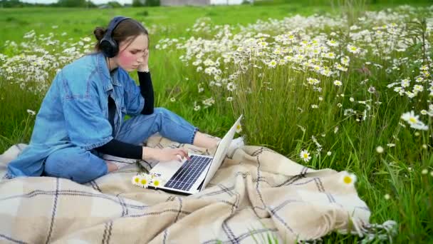 十几岁的女学生坐在公园的草坪上 一边用黑色耳机打电话 一边挥手端着咖啡 一边聊天 穿着休闲裤和蓝色衬裙在空气中工作的女性 — 图库视频影像