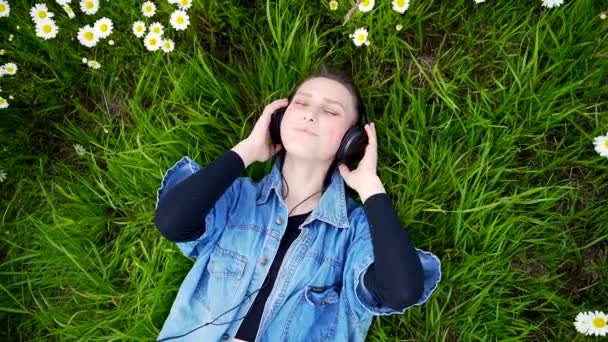 草の中に横たわる若い幸せな笑顔の女の子は晴れた夏の日に大きな黒いヘッドフォンで音楽に耳を傾ける スタイリッシュなティーン大学生とともに茶色の髪のジーンズジャケット公園で芝生の上でリラックス — ストック動画