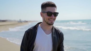 Şık, genç ve çekici bir adamın portresi. Siyah deri ceketli ve güneş gözlüklü bir adam okyanus ya da deniz kenarında mutlu bir şekilde yürür. Güneşli hava, ağır çekim. Seksi beyaz adam sahil boyunca tek başına yürüyor.