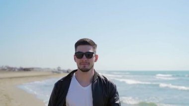  Siyah deri ceketli, güneş gözlüklü, kendine güvenen şık bir adamın portresi. Sakallı bir adam dalgalar ve kumdan oluşan bir arka planda deniz ya da okyanus kıyısında yürür. Güneş, yavaş çekim