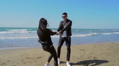 Genç bir aile, aktif olarak el ele tutuşan bir çift. arka planda deniz veya okyanus, el ele gülüşmeler. Samimi ve mutlu bir adam ve siyah deri ceketli bir kız. İyi yolculuklar.