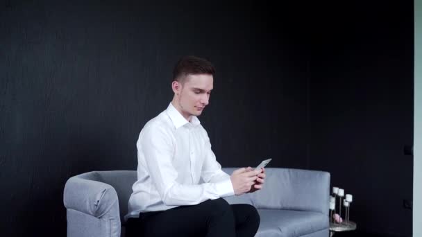 Muž sedí na pohovce a drží telefon. Píše zprávy a čte. Úsměv — Stock video