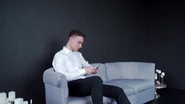 Ein Mann sitzt auf einem Sofa und hält ein Telefon in der Hand. Schreibt Nachrichten und liest. Lächelnd — Stockvideo