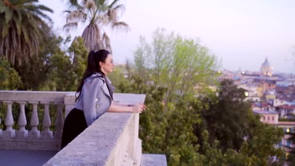 Молодая девушка сидит и стоит рядом с поручнями на фоне старого города. — стоковое видео