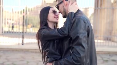 Siyah elbiseli iki gencin portresi. Deri ceket ve güneş gözlüğü. Arka planda Colosseum ve kemer var. Erkek ve kadın turistler el ele tutuşup dans ediyorlar.. 