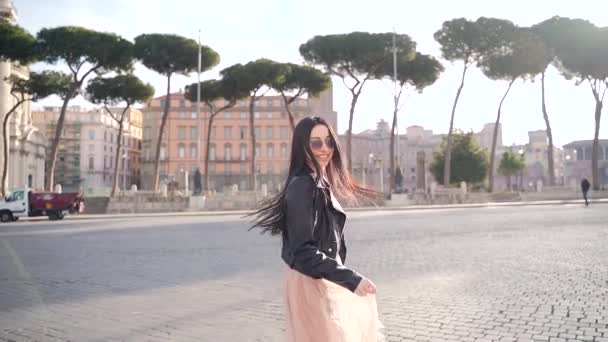 快乐的黑发女孩在相机前摆姿势 在城市建筑背景的鹅卵石广场上微笑和旋转的肖像 意大利 穿着黑色夹克和衣服的游客微笑 — 图库视频影像
