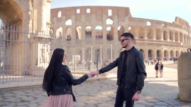 Siyah elbiseli iki gencin portresi. Deri ceket ve güneş gözlüğü. Arka planda Colosseum ve kemer var. Erkek ve kadın turistler el ele tutuşup dans ediyorlar.. 
