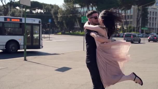 在城市背景下遇见一对快乐的夫妇 城市广场 公路交通 穿着裙子的女孩奔向那个拥抱她脖子 纺纱和亲吻的男人 道路和汽车 汽车站 慢动作 — 图库视频影像