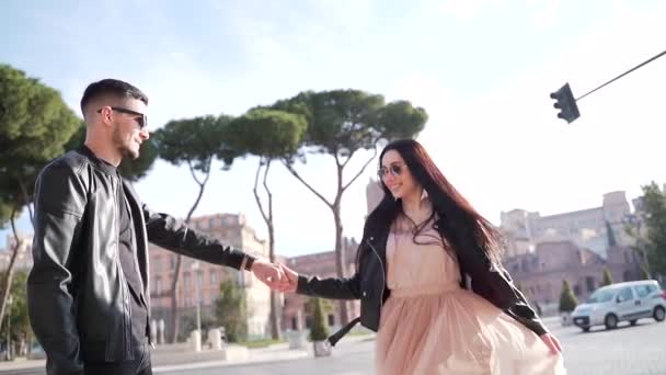 年轻快乐的一对男女手牵着手拥抱跳舞 开心点男人和女人穿着时髦的衣服 背景是古城的树木和靠近长椅的建筑 意大利罗马 — 图库视频影像
