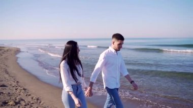 Kot pantolon ve beyaz gömlekli genç çift deniz kenarında yürüyor. Genç bir kadın ve erkek el ele tutuşup sahil boyunca yürüyor. Kum, okyanus ve dalgalar arka planda. Mutlu kız ve erkek sarılıyor 