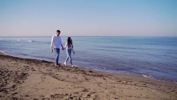 年轻夫妇穿着牛仔裤和白衬衫在海边散步 年轻的女人和男人牵着手沿着海岸走着 背景上的沙子 海洋和波浪 快乐的女孩和男人拥抱 — 图库视频影像