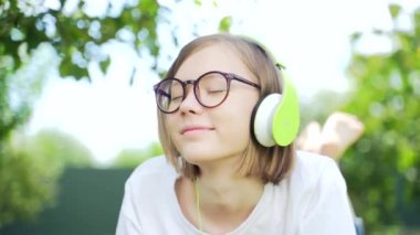 Parkta ya da bahçede kulaklıkla müzik dinleyen genç bir kızın yakın plan portresi. Çekici hippi sarışın, gözlüklü ve beyaz tişörtlü, gözleri kapalı, doğada rahatlayan. dinlenme