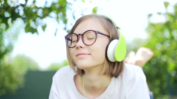 在公园或花园用耳机听音乐的少女的特写 迷人的嬉皮士金发碧眼 戴着眼镜 穿着白色T恤 闭着眼睛 本质上很放松 休息一下 — 图库视频影像