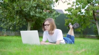Genç bir kadın çimlerde dizüstü bilgisayarla yatıyor. Parktaki çim üzerinde çalışan ve ders çalışan kız öğrenci. Güneşli bir günde beyaz tişörtlü ve kot pantolonlu bir kadın. Mesafe öğrenme. Doğada rahatla.