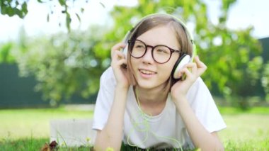  Parkta ya da bahçede kulaklıkla müzik dinleyen genç bir kızın yakın plan portresi. Çekici hippi sarışın, gözlüklü ve beyaz tişörtlü, gözleri kapalı, doğada rahatlayan. dinlenme