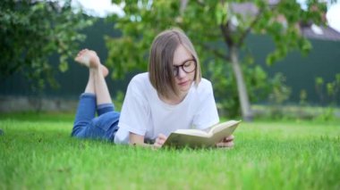 Öğrenci kız parkta çimlerin üzerinde bir kitap okuyor. Genç bir kız çimlerin üzerinde uzanıyor ve elinde bir kitap tutuyor. Gözlüklü bir kadın. Sarışın, beyaz tişört ve kot pantolonla ders çalışıyor. Yeşil çimen, güneş, yaz
