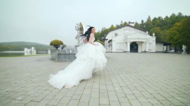 Moda gelini koşar ve kameraya bakar. Parlak gelinlikli esmer bir kadın uzun trenli güneşli bir günde çeşmenin yanında kaçar. Yavaş çekim Arka tam uzunluk görünümü