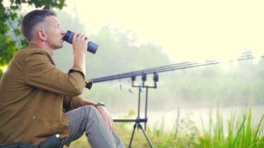 Balıkçı portresi balıkçısı sandalyede oturmuş, bir şeyler içiyor, termostan çay içiyor. Göl oltalarının ve ağaçların arka planında doğa var. Hipster adam tatilde. Etkin yeniden canlandırma kapat