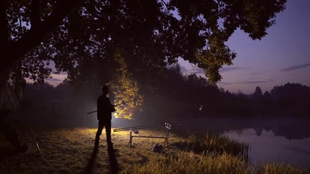 シルエットは男だ 若い男の漁師が湖のそばに釣り竿を持って立っている 男はリールを持った魚を捕まえる 水の上に夜や朝霧 魚のタックル キャンプやお気に入りの趣味 — ストック動画
