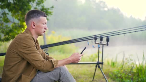 年轻英俊迷人的渔夫坐在湖边或河边的椅子上 手里拿着智能手机 一个天生带着电话的家伙鱼的背景是杂物和钓竿 人物形象 — 图库视频影像