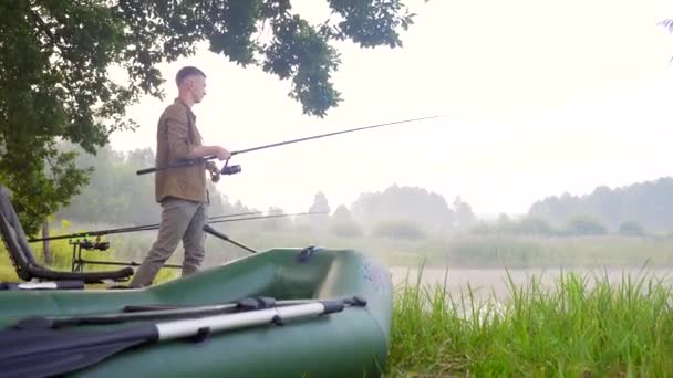 漁師は湖のそばで回転するか釣竿で立っている リールを持ってる 膨脹可能なボート 朝霧葦の木 装備魚が取り組む キャンプや趣味 ヒップスターの肖像 — ストック動画