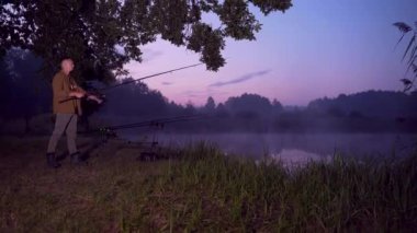 Siluet bir adam. Genç erkek balıkçı elinde dönen bir oltayla gölün kenarında duruyor. Adam makaralı bir balık yakalıyor. Gece ya da gündüz sisi, balıklar kapışıyor. Kamp ve favori hobi